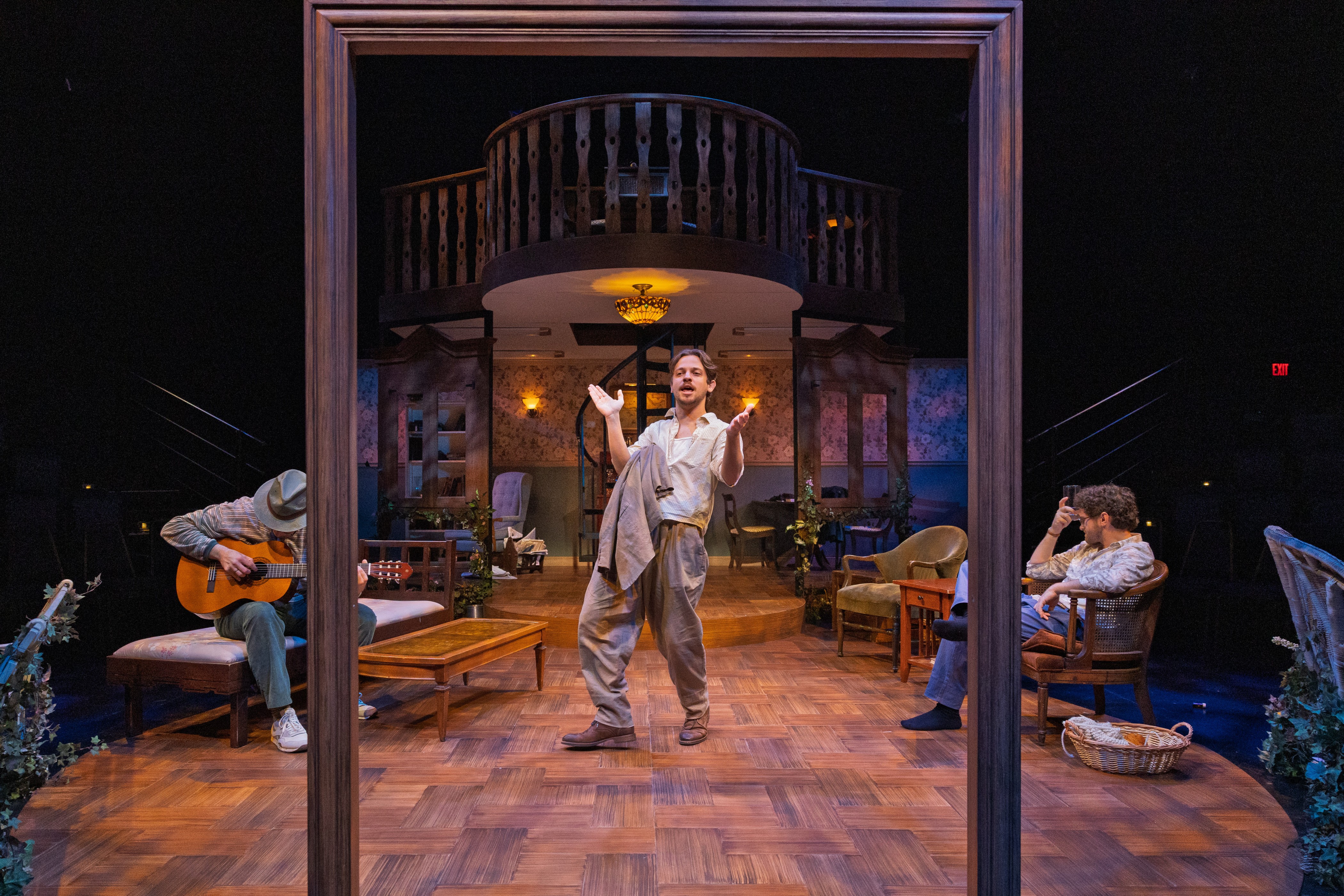 A theatrical production shot of Uncle Vanya showing three performers on a warmly lit stage. Center, a man in a rumpled white shirt and loose trousers stands with arms raised and open, mid-speech. To the left, a performer in a wide-brimmed hat sits playing an acoustic guitar. To the right, another performer sits in a chair looking at a phone. The set depicts a lived-in interior with floral wallpaper, vintage furniture, potted plants, and a curved staircase leading to an upper balcony, all framed by a large wooden doorframe in the foreground.