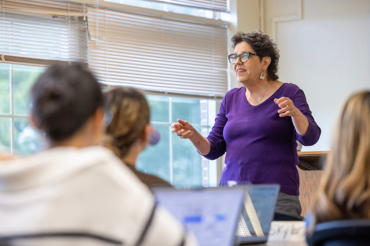 A female teacher stands in front of sitting students speaking and gesturing with her hands