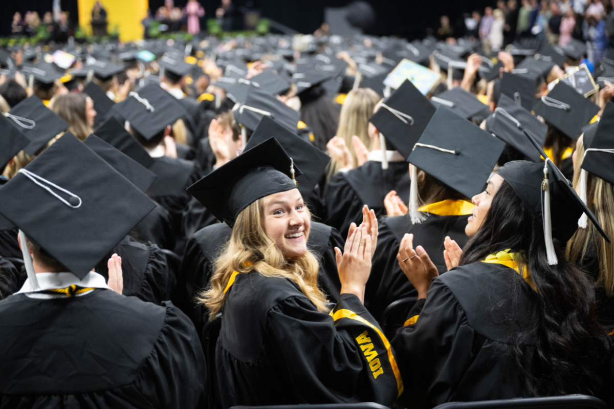 A graduating student among several in regalia looks behind herself as she claps, smiling