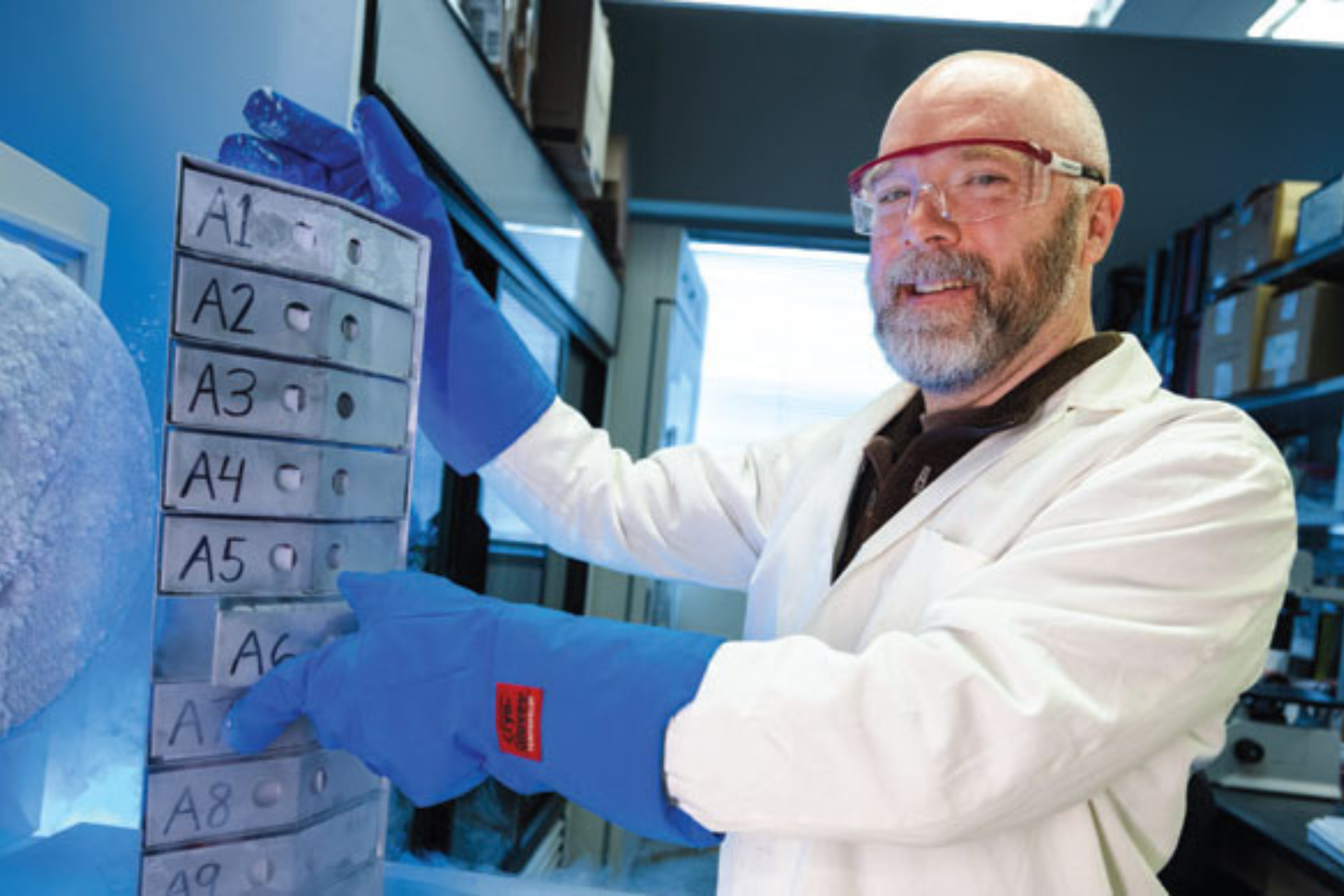 Brian Berger stands in lab gear, points to a sign in blue gloves.