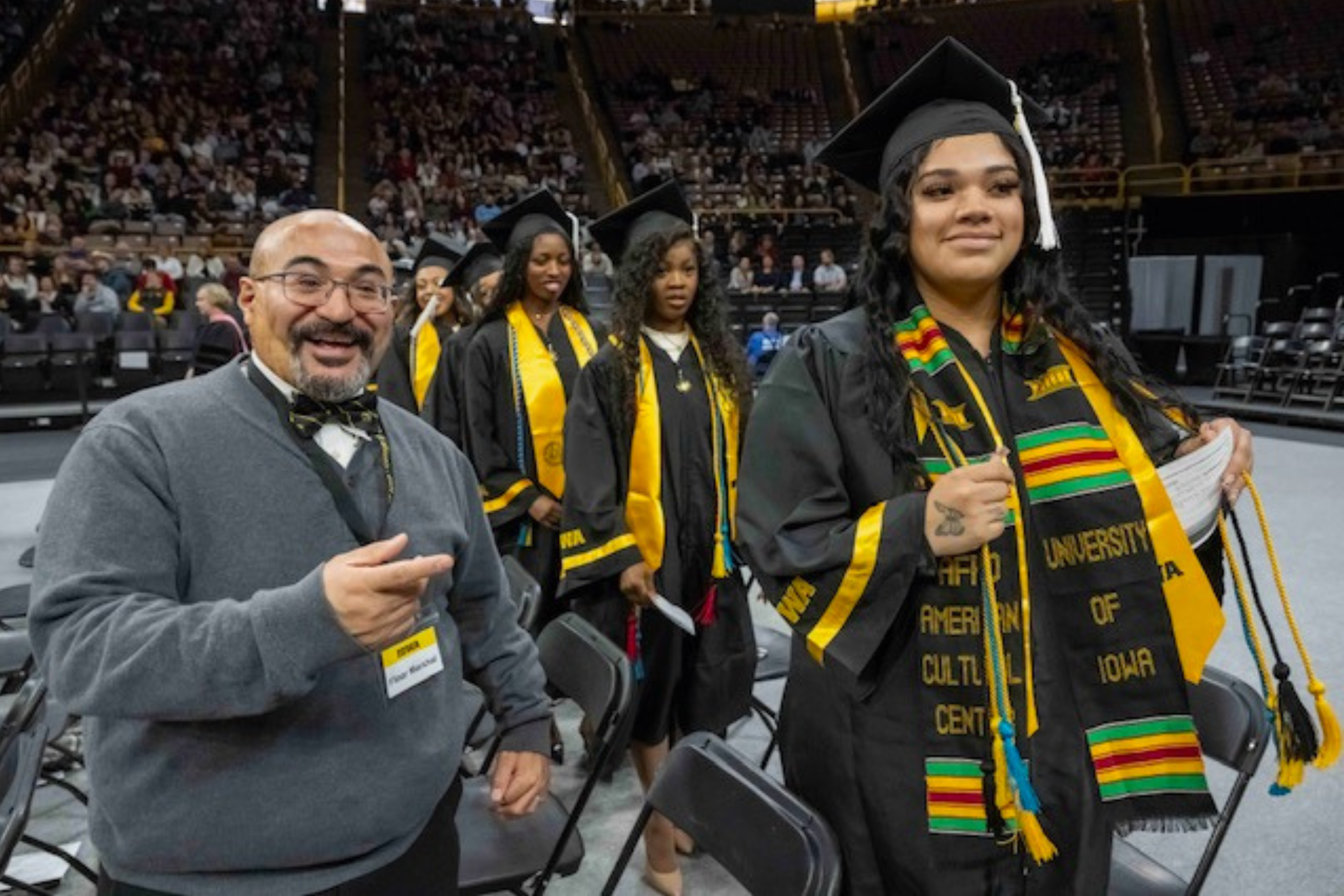 a woman amongst a crowd of fellow graduates in cap in gown smiles and waves to someone in the stands