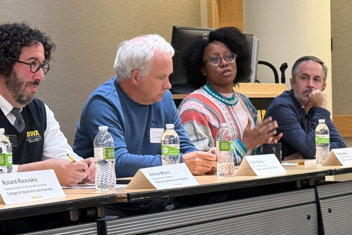 Four panelists with nameplates in front of them face a crowd, one is speaking and gesturing with her hands