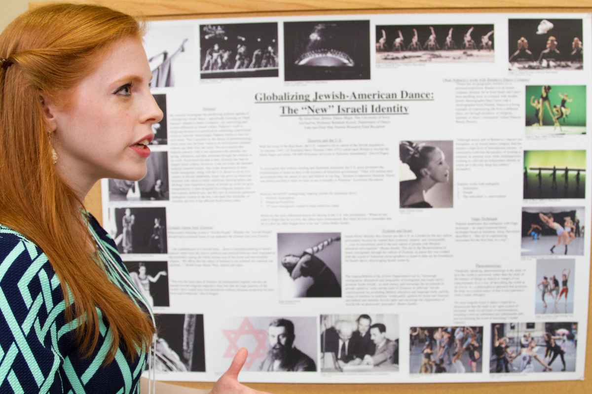 A woman is in profile talking to someone off-camera. A poster with research findings is behind her.