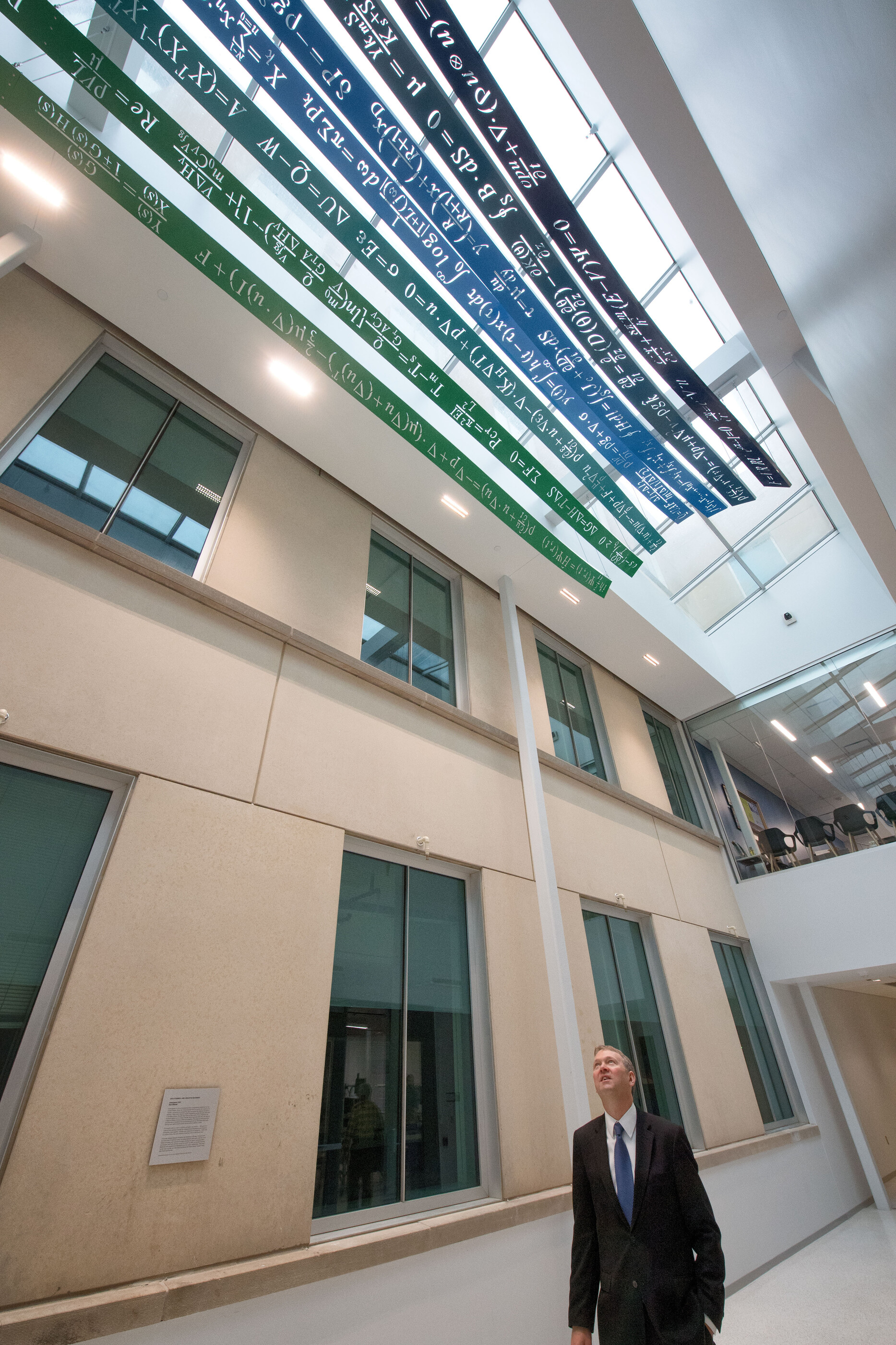 Man stands in corridor looking up at sculpture containing six seperate ribbons hanging parallel 