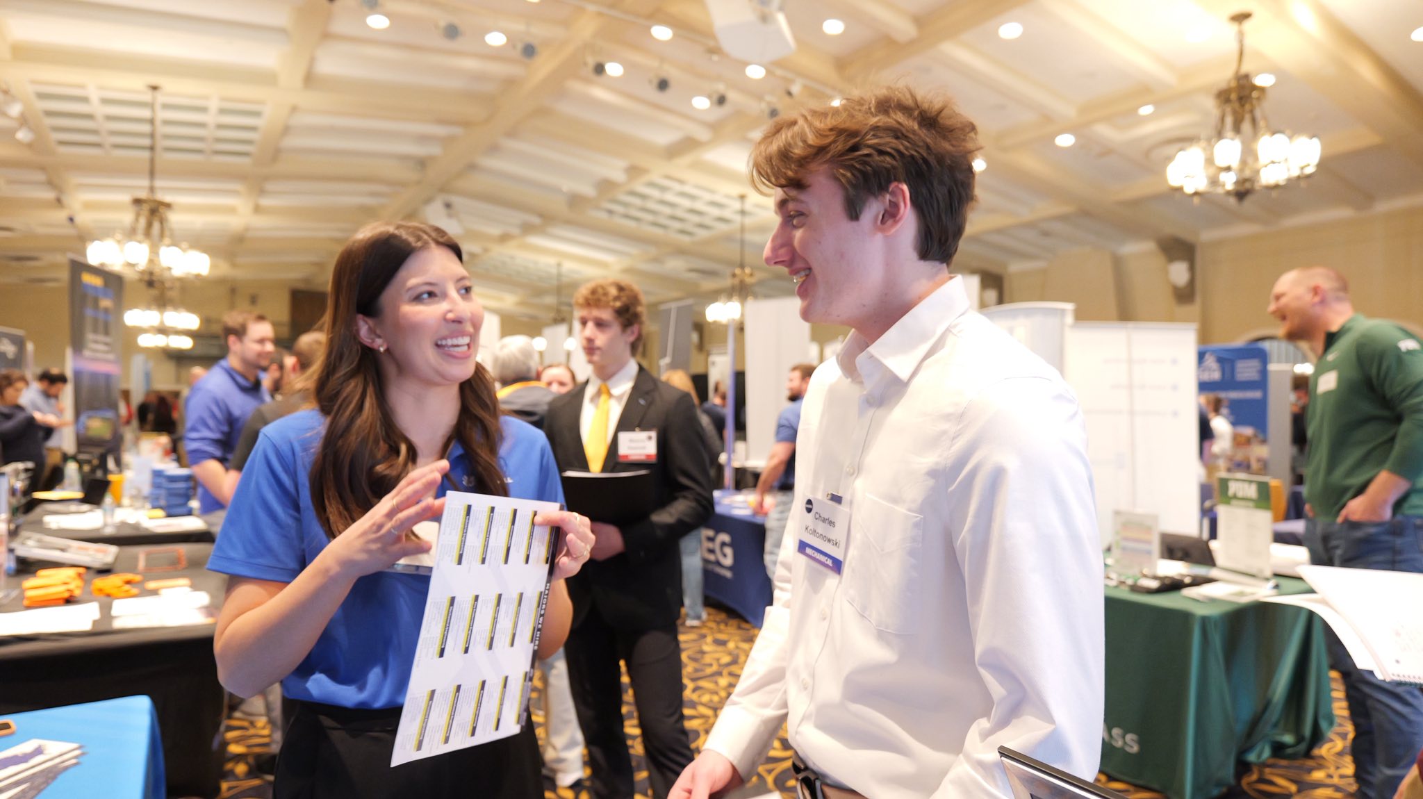 Young man (right) speaks with young professional woman (left) in large room.