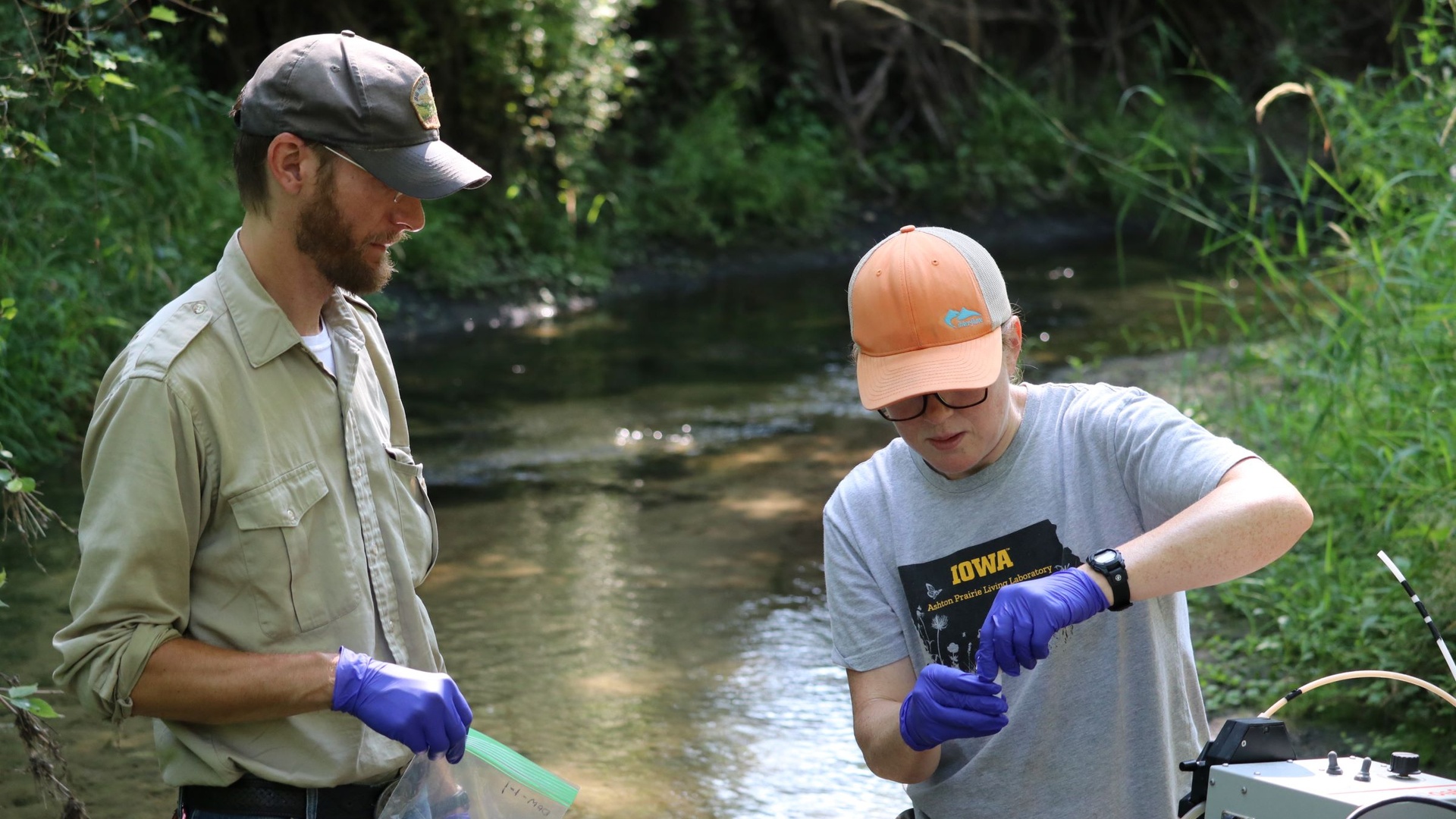 Man (left) and student with purple gloves stand by a waterway reviewing a sample