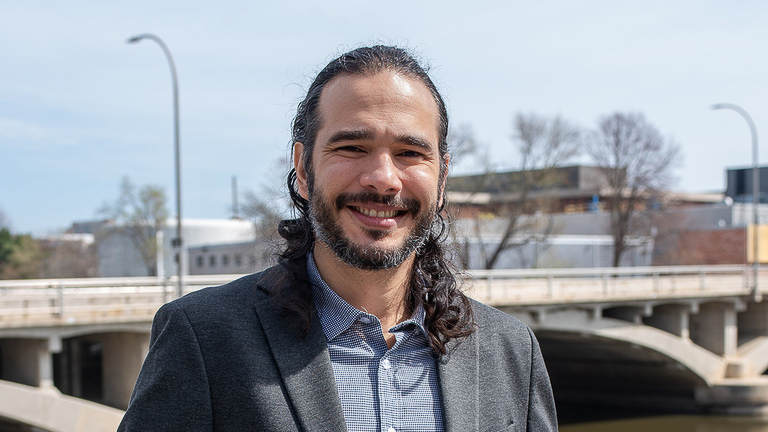 Man with long hair and bear stands in front of a river