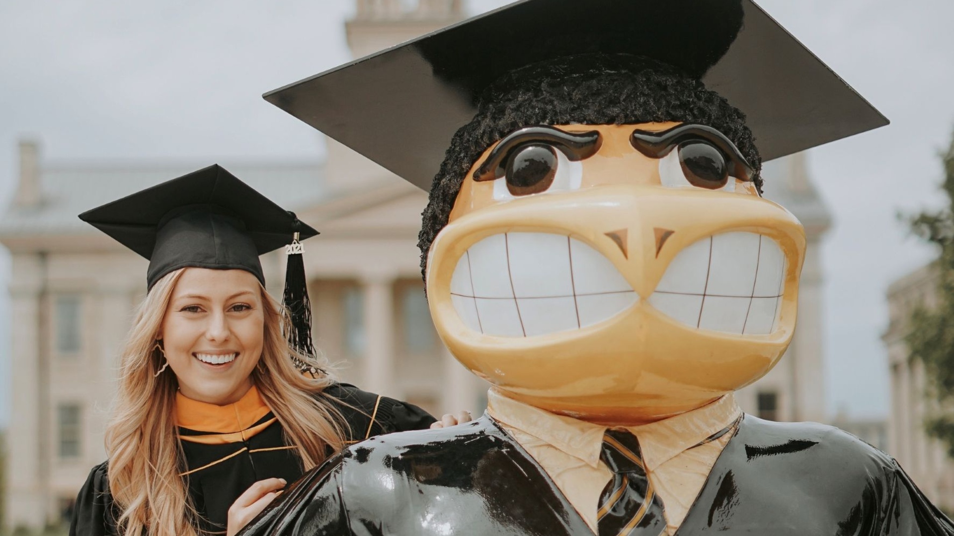 Woman with blond hair wears graduation regalia while posing next to a statue of the Iowa mascot also wearing graduation regalia