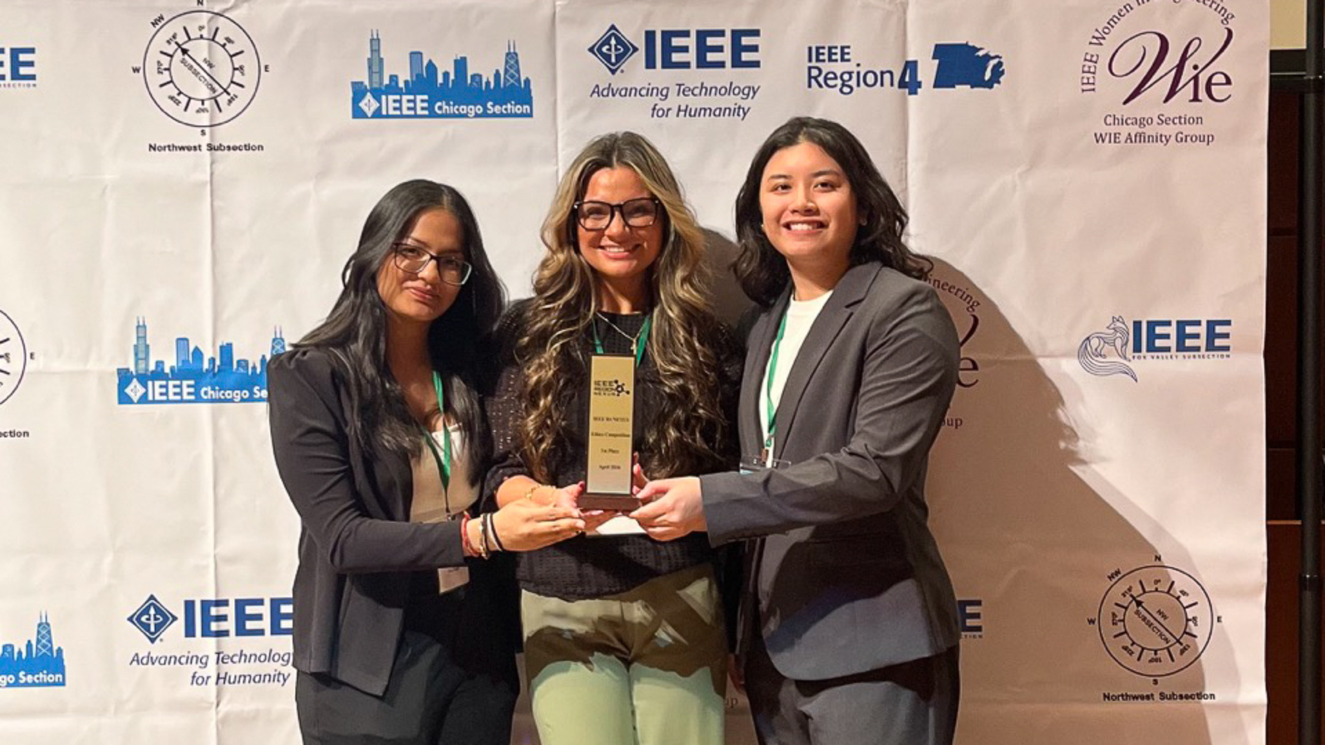 Three college-aged women wearing professional clothing pose with a trophy