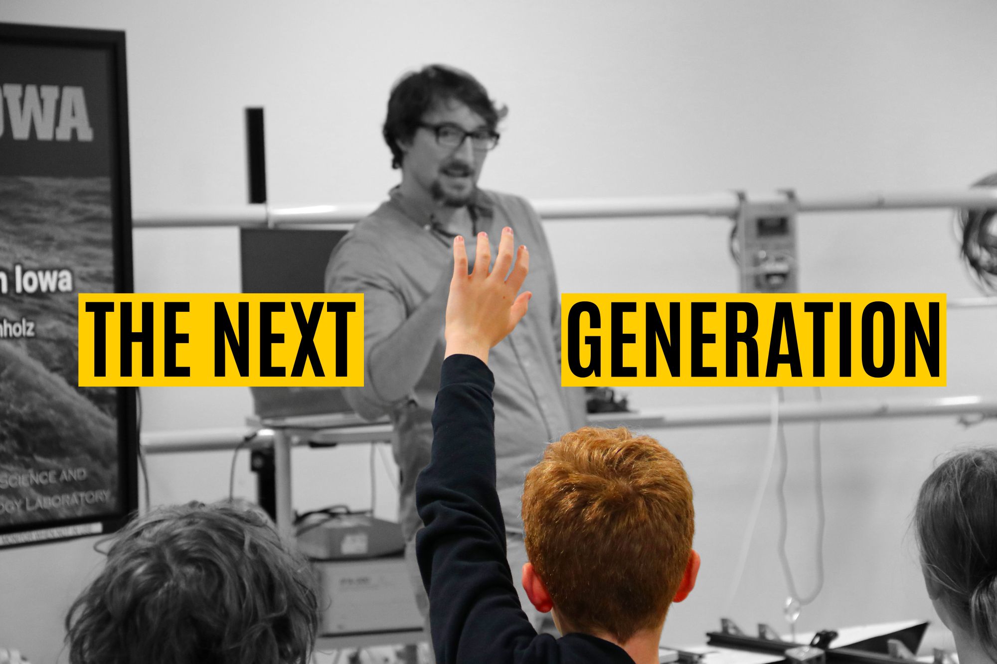 An elementary aged boy raises his hand, with College of Engineering professor Casey Harwood in the background pointing to him