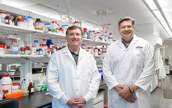Michael Schnieders and Lewis Stevens stand in the Stevens lab in the UI College of Pharmacy, standing in front of rows of medicine bottles