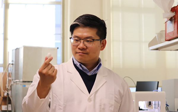 Xuan Mu stands in his lab holding a small white silkworm cocoon pellet about 1 inch tall, used in his research