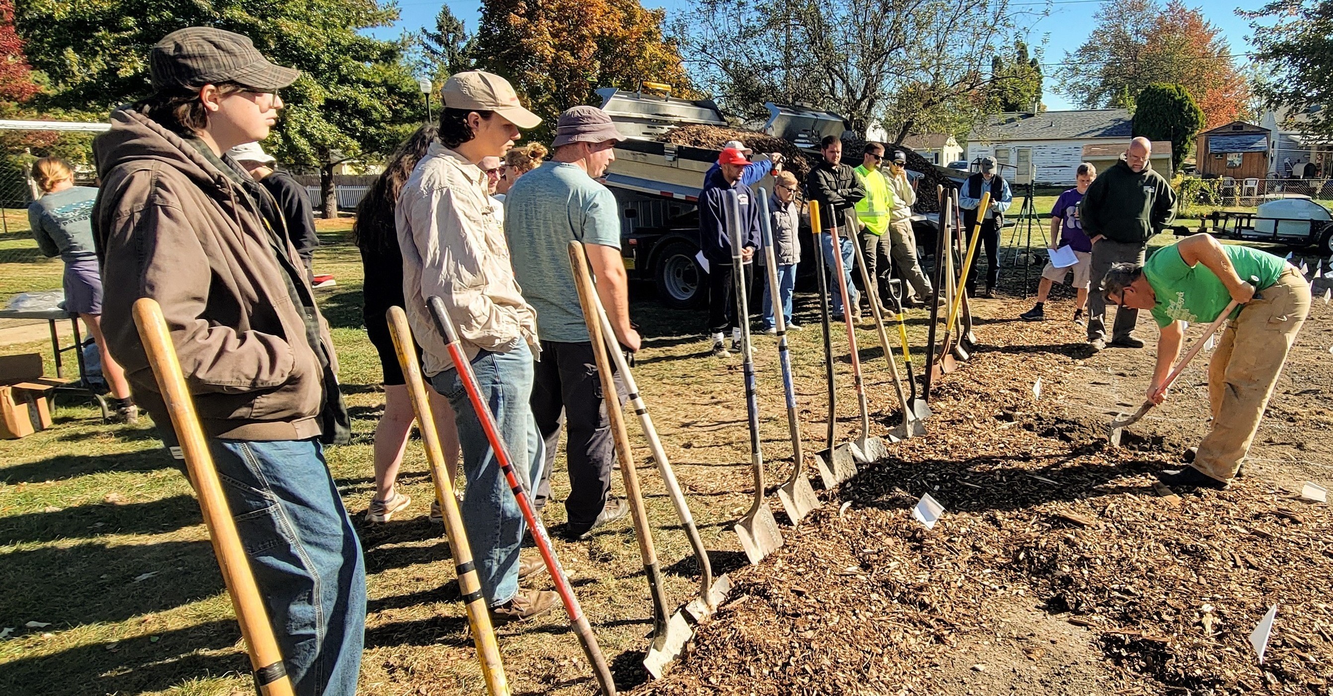 A group of people watches a man digging in the dirt with a shovel surrounded by small flags and tools.