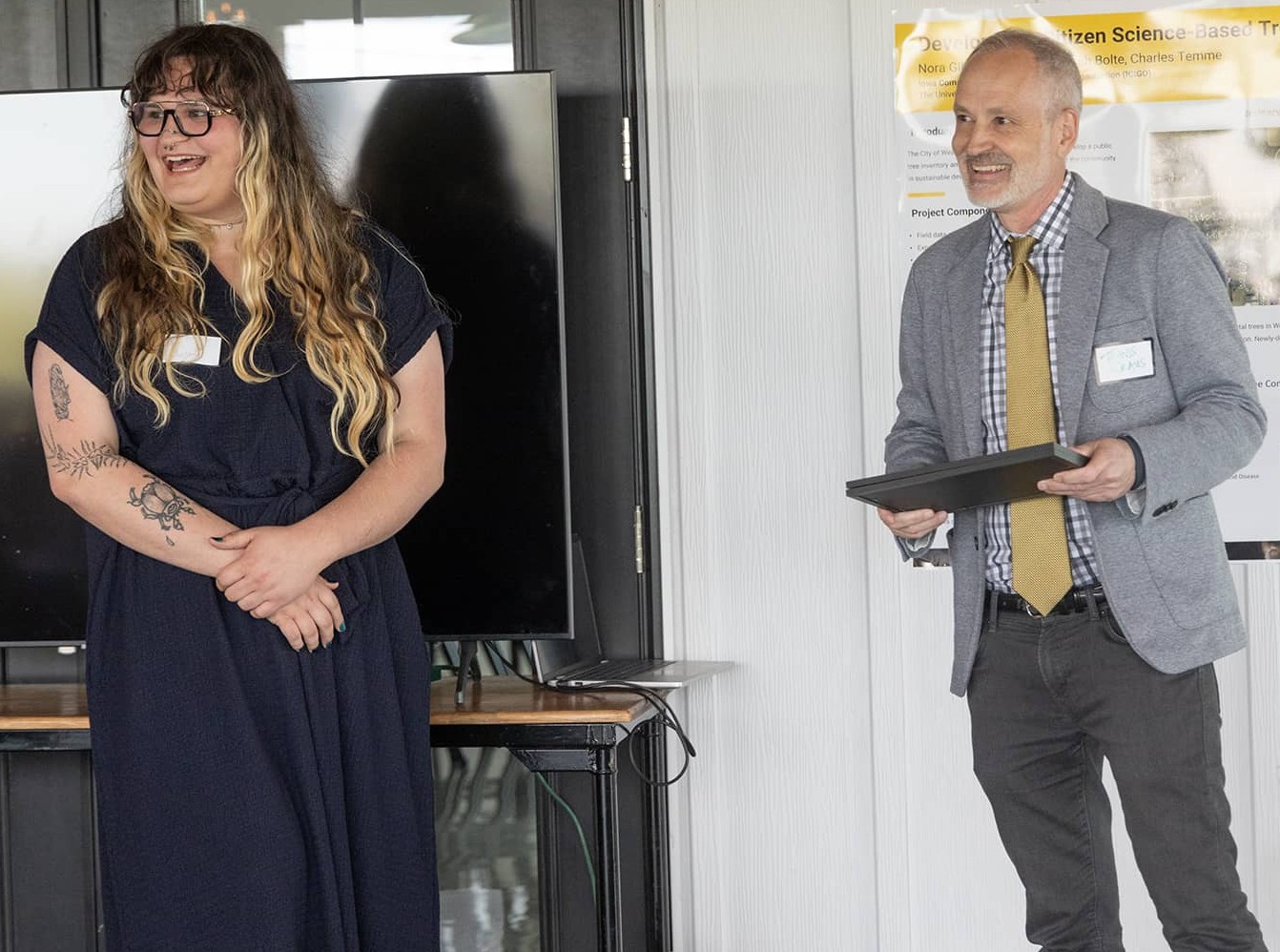 two people stand smiling. The man on the right is holding an award certificate.