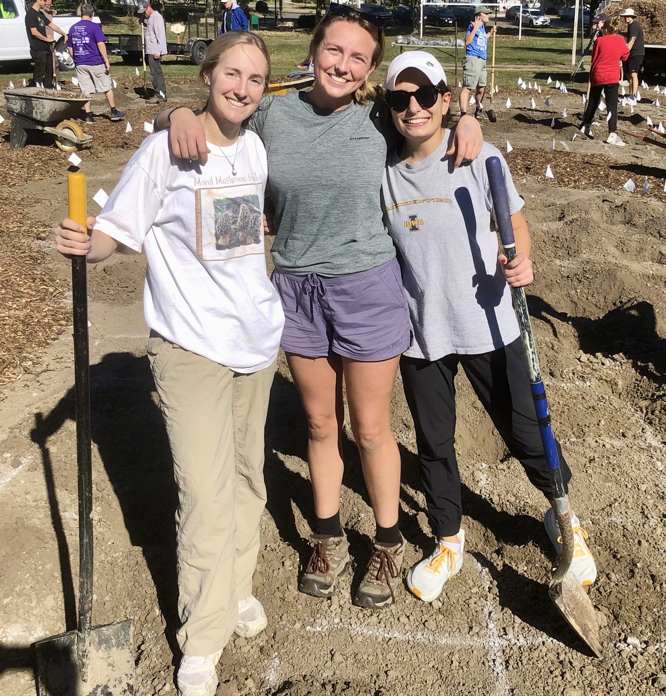 Three women holding shoves and smiling at a worksite with dirt 