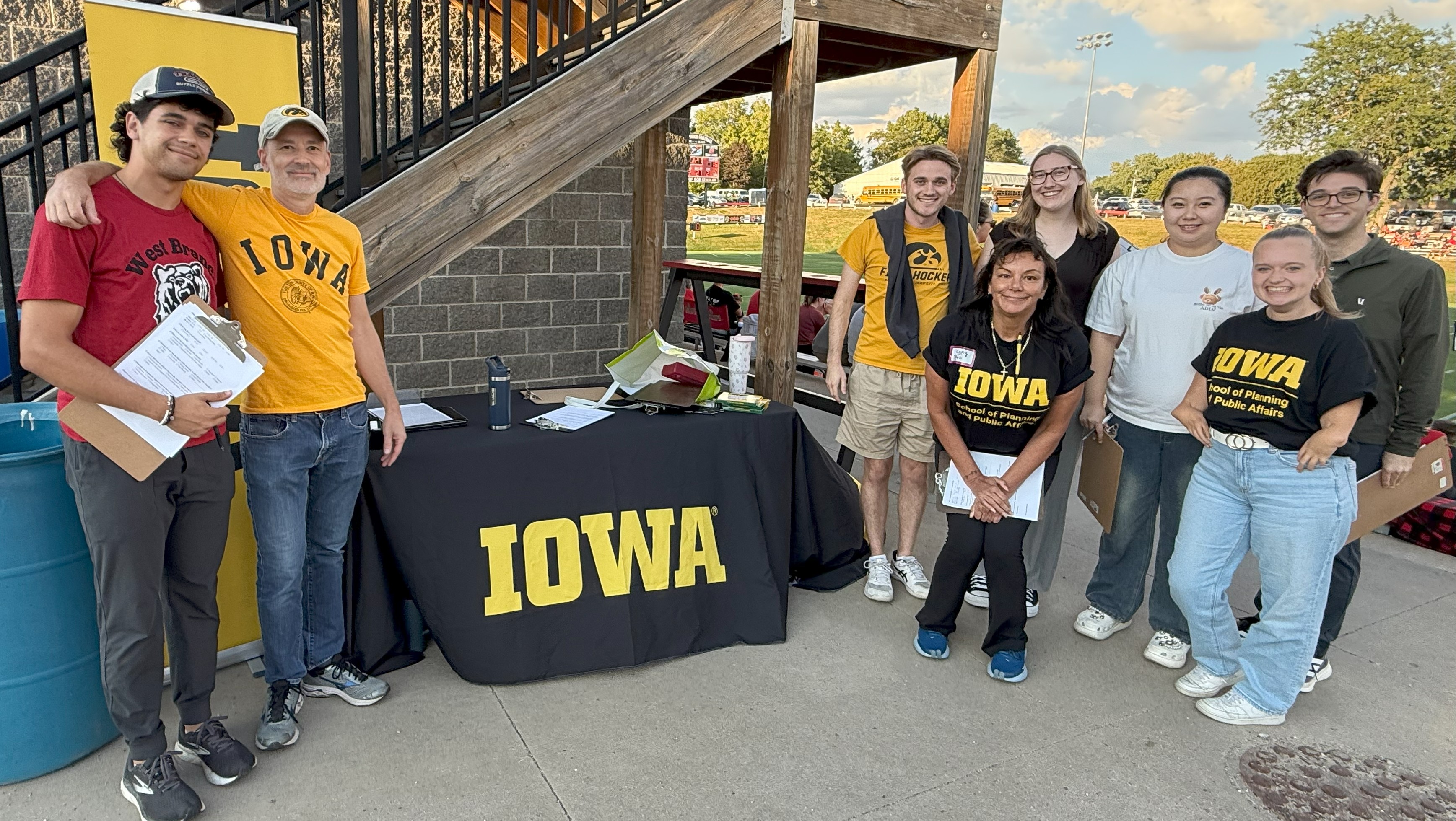 A group of University of Iowa student stand around a table with an Iowa banner at a football stadium