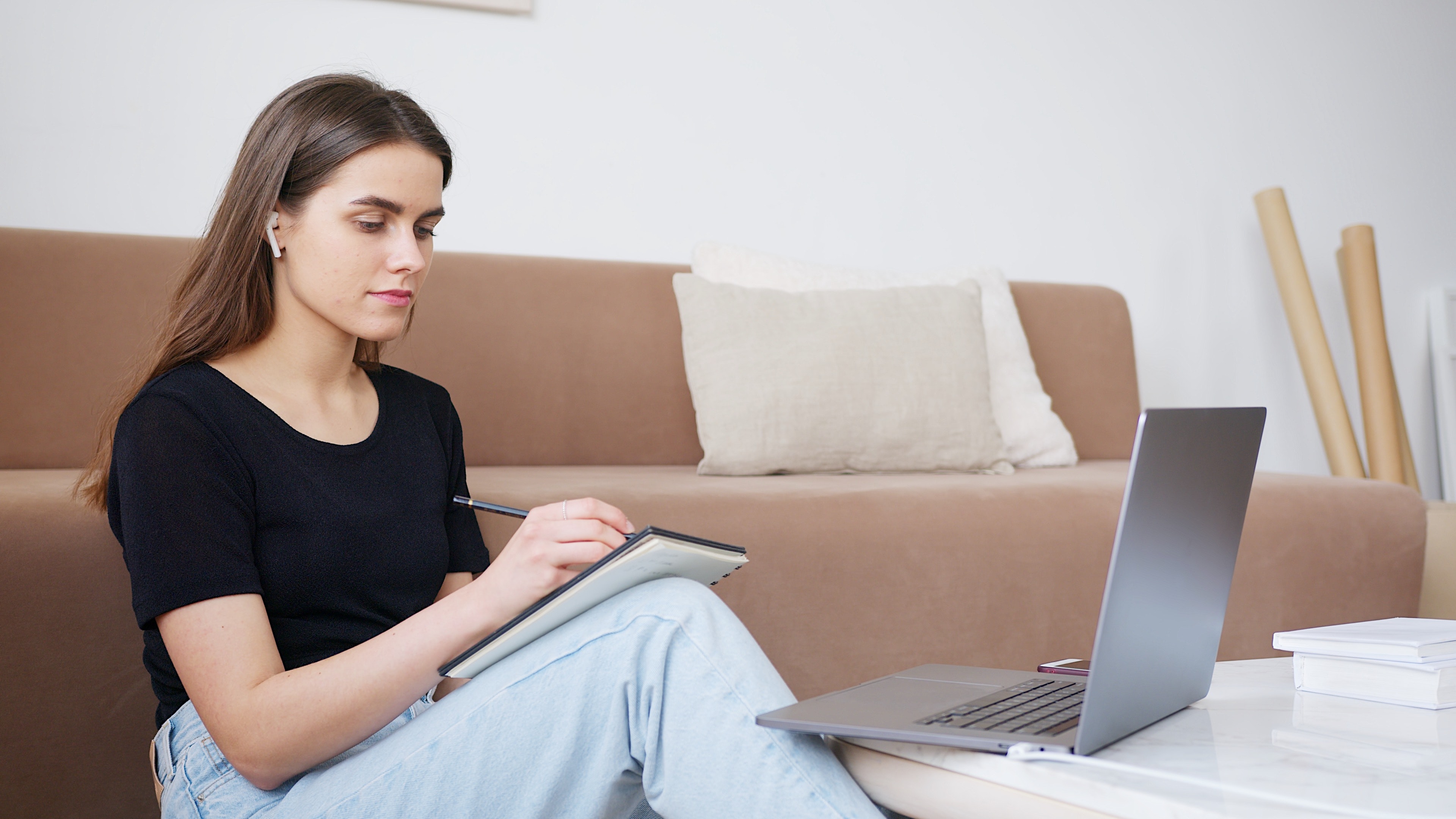 College student sitting on the floor watching their laptop and taking notes in a notebook while sitting on the floor. 