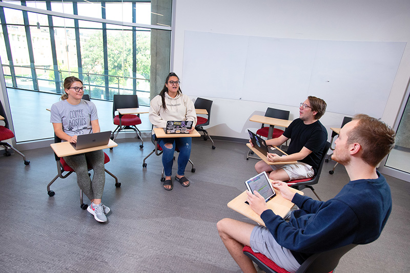 Four University of Iowa students with their desks in a circle while talking with their laptops open. 
