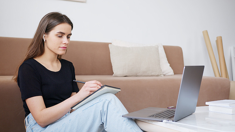 Person sitting on the floor with a notebook while looking at a laptop screen.