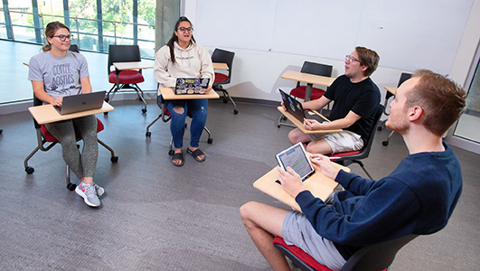 UI students sit in desks while discussing in a classroom.
