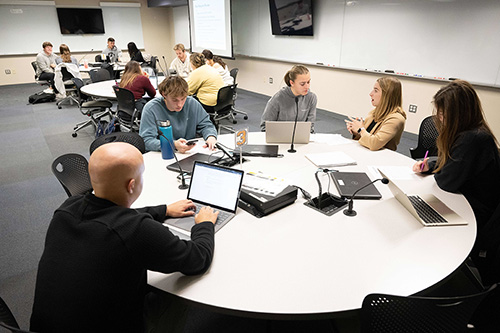 UI students working in a large TILE classroom filled with circular tables, screens on the walls, and laptops.