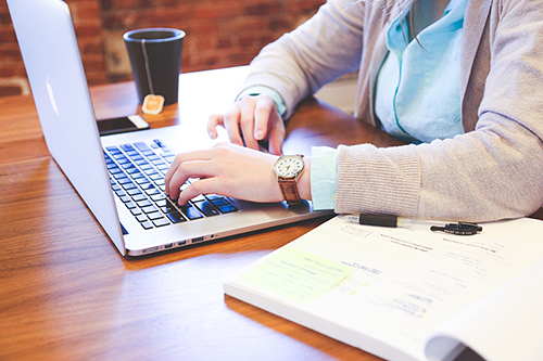 Close up of someone typing on a laptop while an open notebook sits on the table.