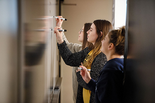 Three UI students writing on a whiteboard in a classroom.