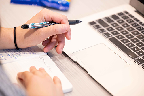 Closeup of a student's hand writing on a note book and using a laptop.