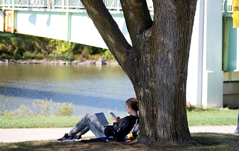 Student sits on green space using their phone and laptop.