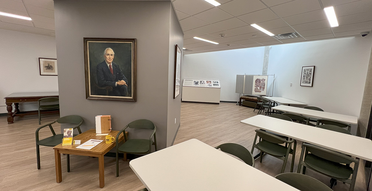 Color photo of the recentl renovated JMRBR, showing a portrait of Dr. John Martin hanging on a gray square column in the middle of the picture, a small portrait of Dr. William Osler hanging over his former desk made from a dark wood, an exhibit about Dr. Martin in the back of the room, the unbound Mascagni (1828) anatomy exhibit to the right of the Dr. Martin Exhibit, and four white tables with green woodden chairs arrayed on the right side fo the photo.