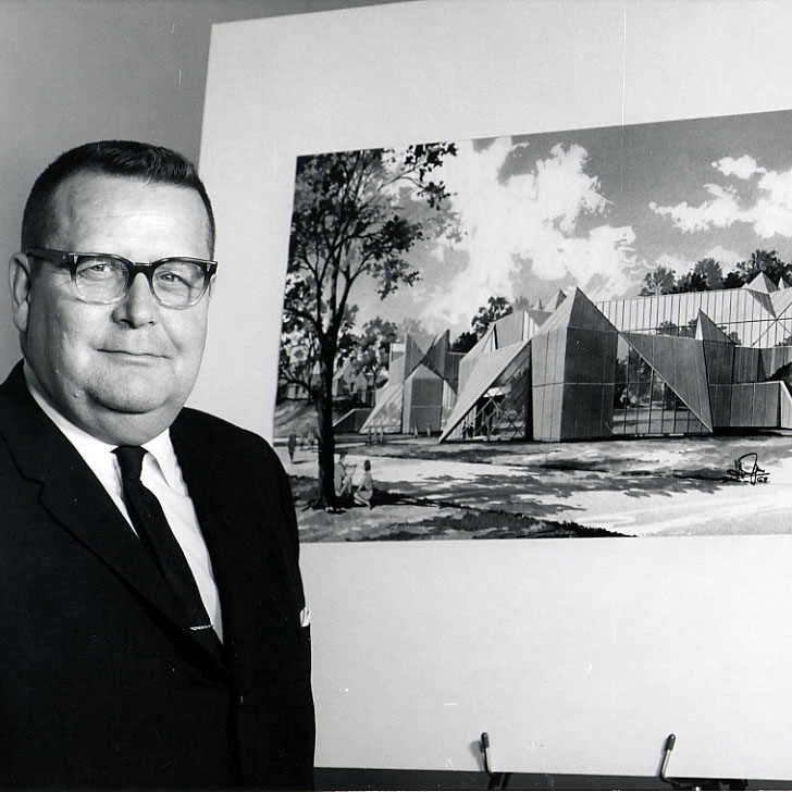 Large white man in a dark suit and tie standing next to an artist's rendering of the planned Hardin Library in 1970.