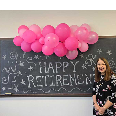 A color photo of a retirement celebration featuring a woman in a dark dress with a pink and white floral print standing in front of a blackboard with Happy Retirement written on it in chalk and a collection of pink balloons hanging down from the top of the board.