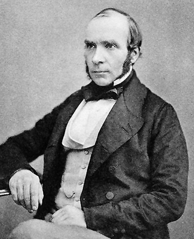 Black and white photograph showing a white man with sideburns, a black jacket, white shirt, and a bow tie, sitting in a chair next to a desk.
