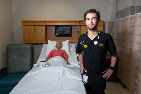 Grayson Wills in nurse's scrubs with a stethoscope around his neck. He's standing beside a hospital bed that contains a pediatric simulated patient.