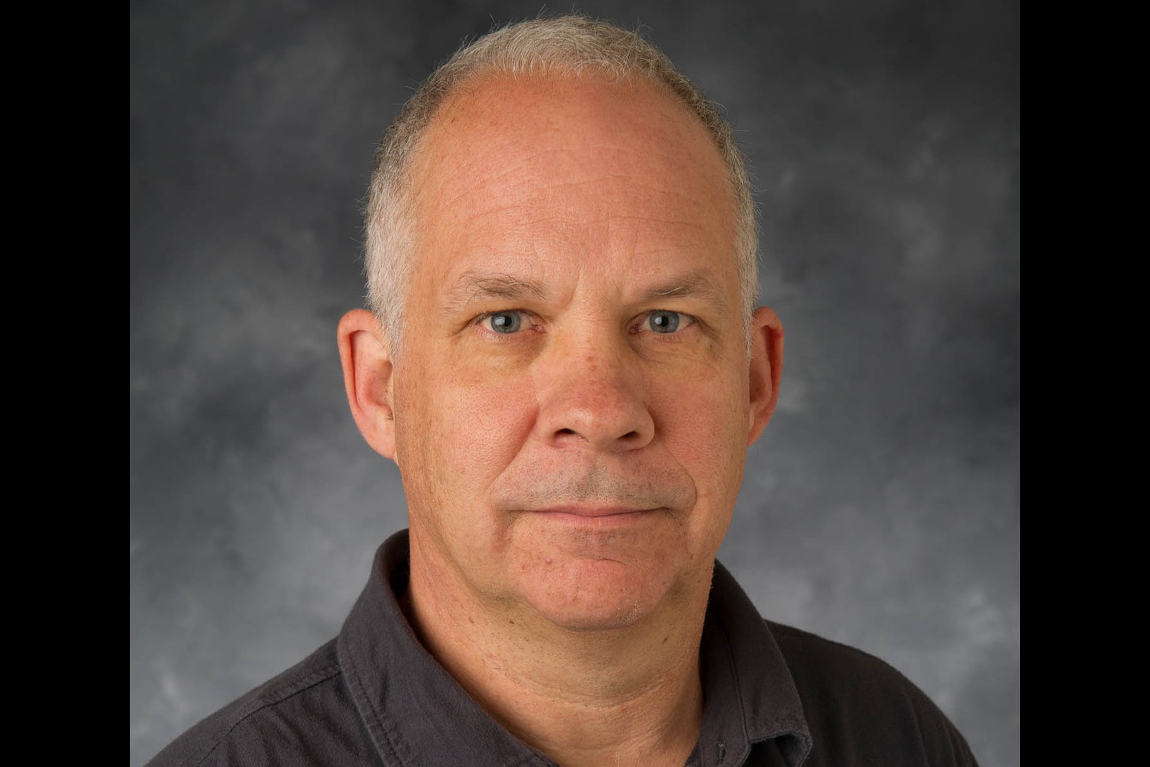 A close-up of Colin Gordon, a man with close cropped salt and pepper hair, wearing a black polo shirt.