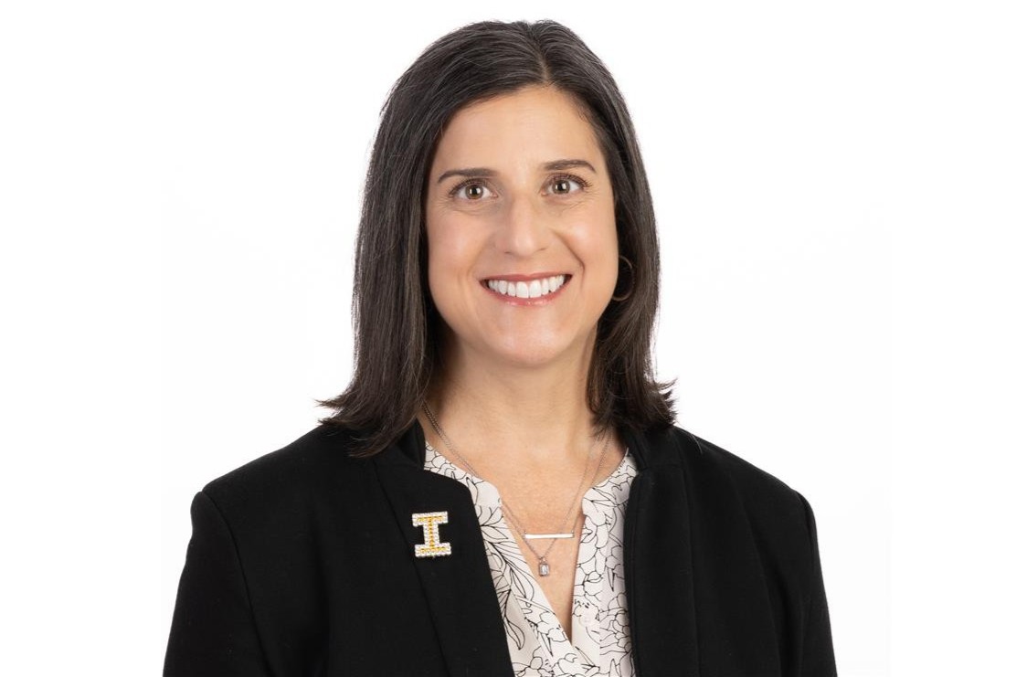 Sheila Baldwin has mid-length dark hair, is smiling, and wearing a dark suit jacket with a sparkling yellow I pin, and a white and black blouse.,