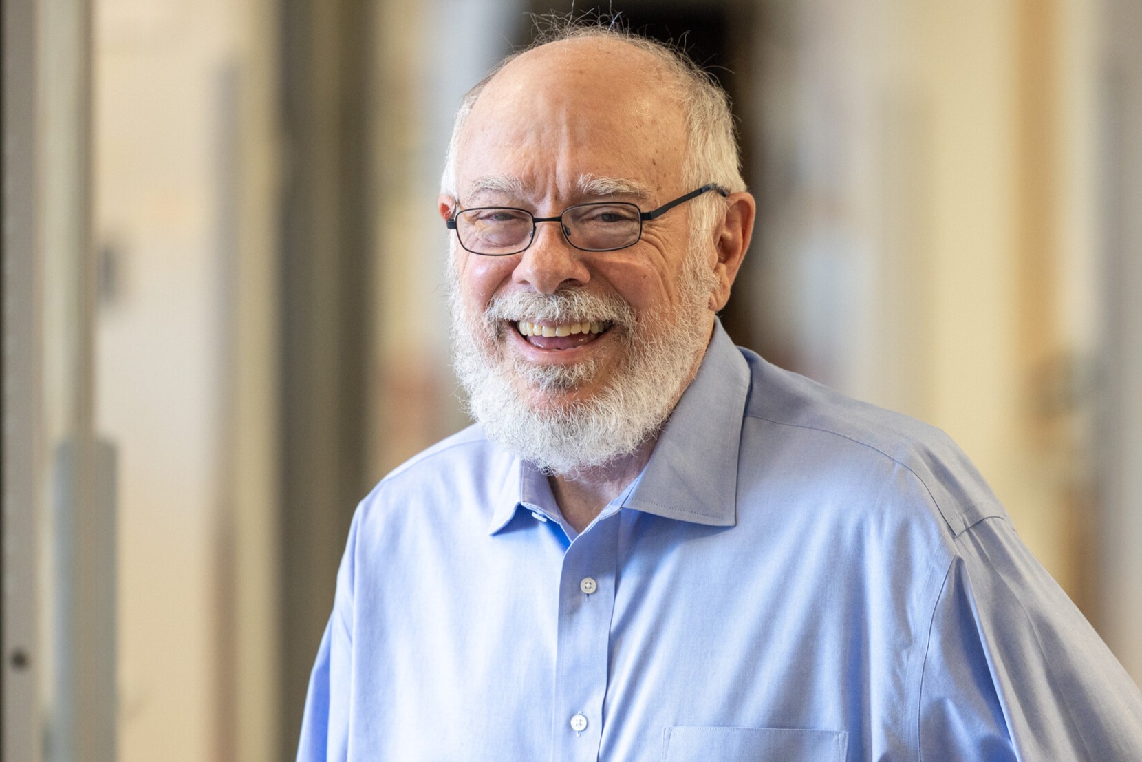 Kevin Campell, is a man with a beard, silver rectangular glasses, wearing a blue button down shirt. He appears to be standing in a laboratory setting.