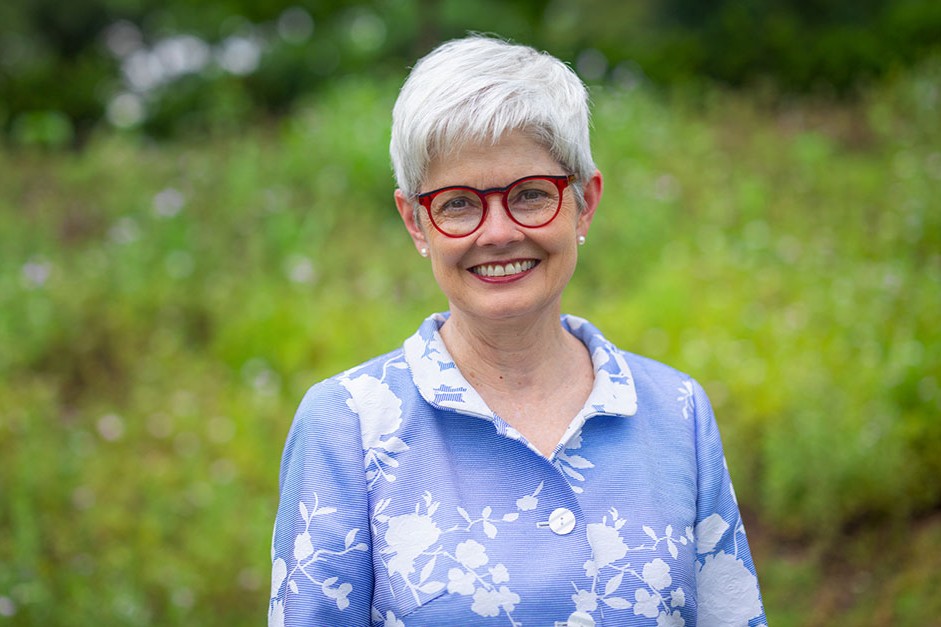 Lynette Marshall, a woman with short white hair, red glasses, wearing a blue and white floral blouse. She appears to be standing in a garden. 