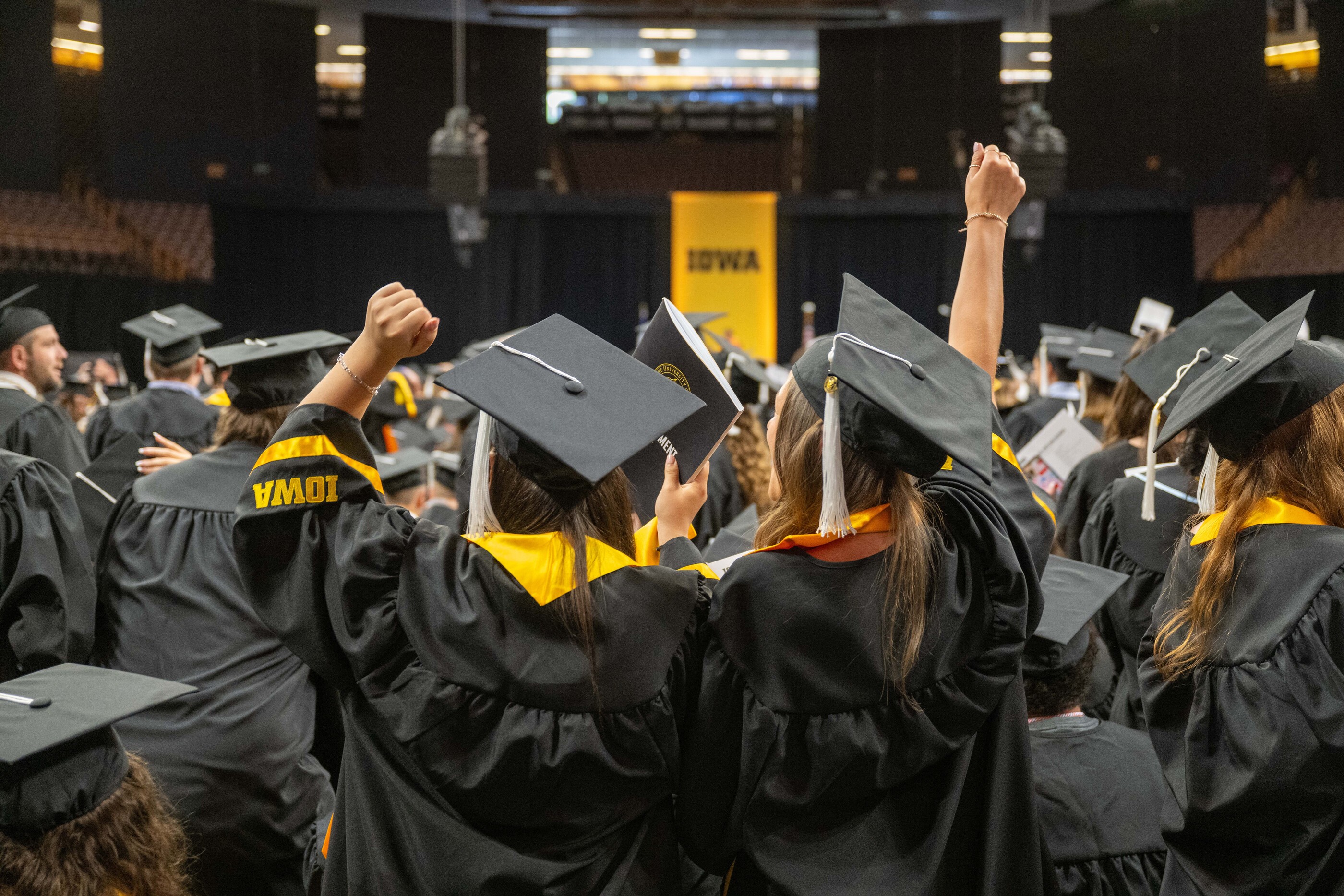 Two students with their arms up in triumph at the commencement ceremony. They are facing the podium and are surrounded by other students in caps and gowns.