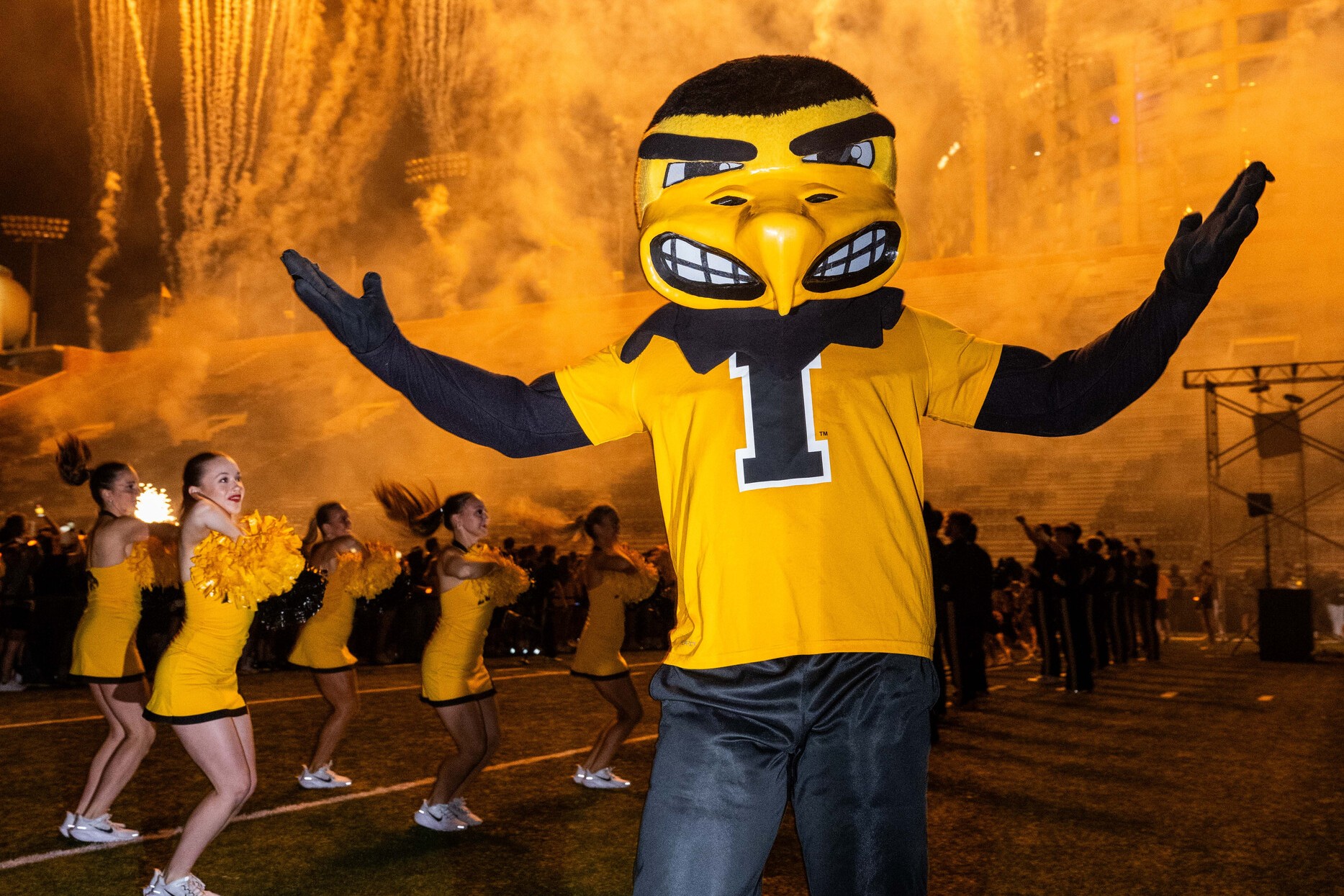 Herky the Hawk standing on the football field with cheerleaders and fireworks in the background.