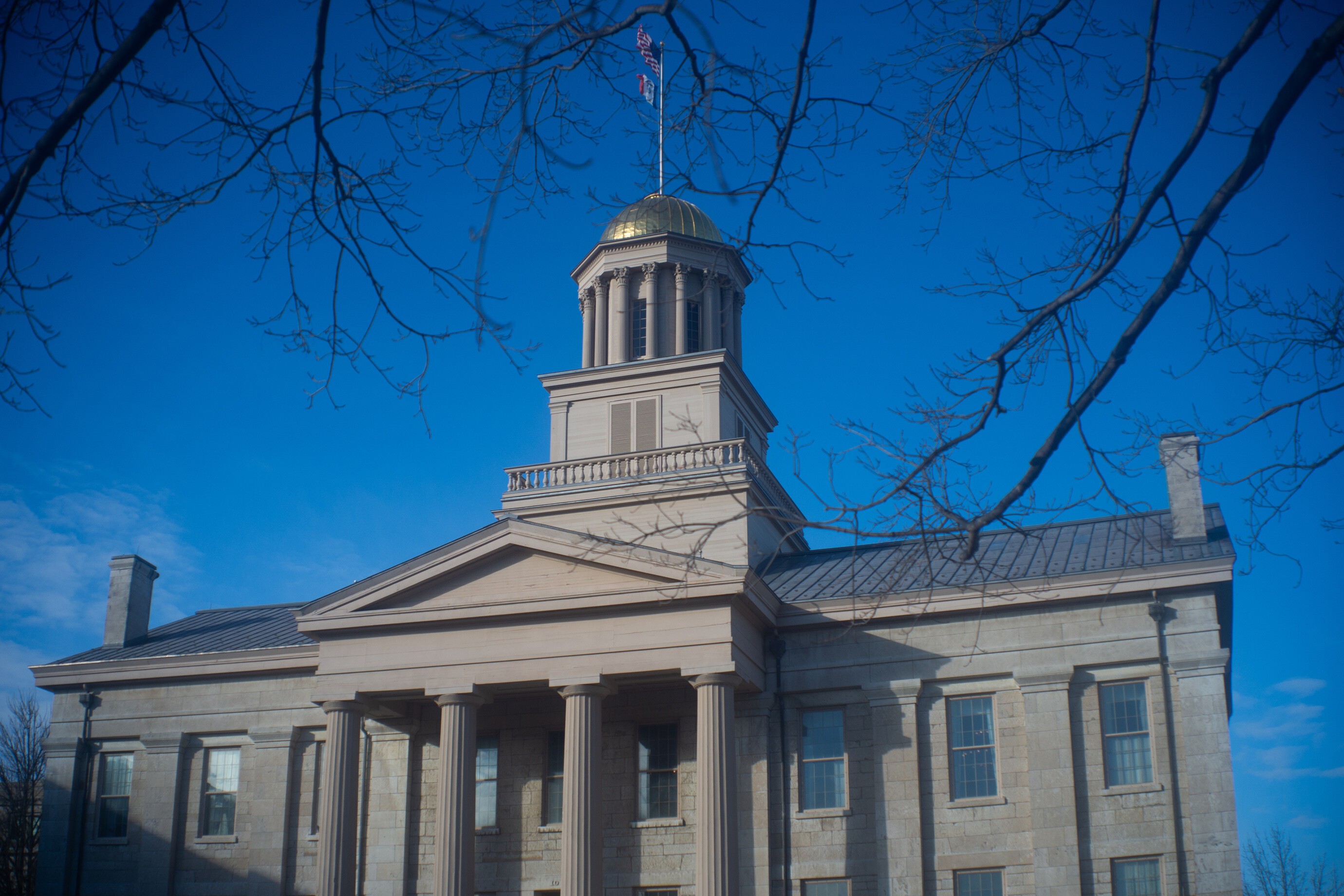 Old Capitol against a deep blue winter sky, framed by leafless tree branches. The gold dome gleams in the bright sunlight.