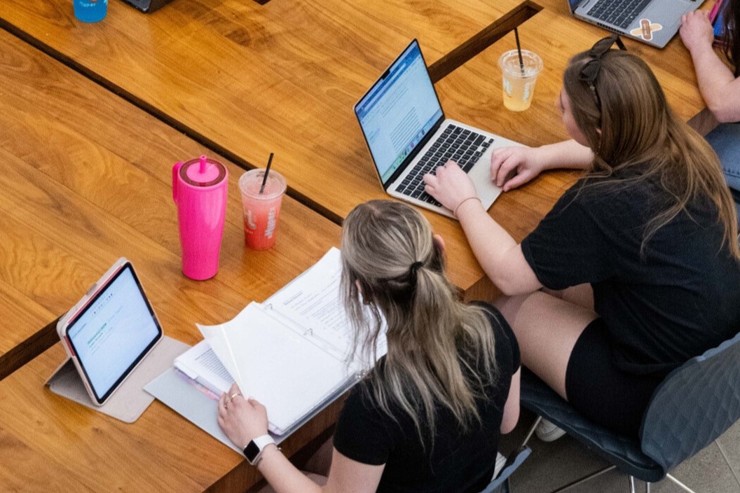 An overhead view of two women studying at a table with their laptop and tablet open. 