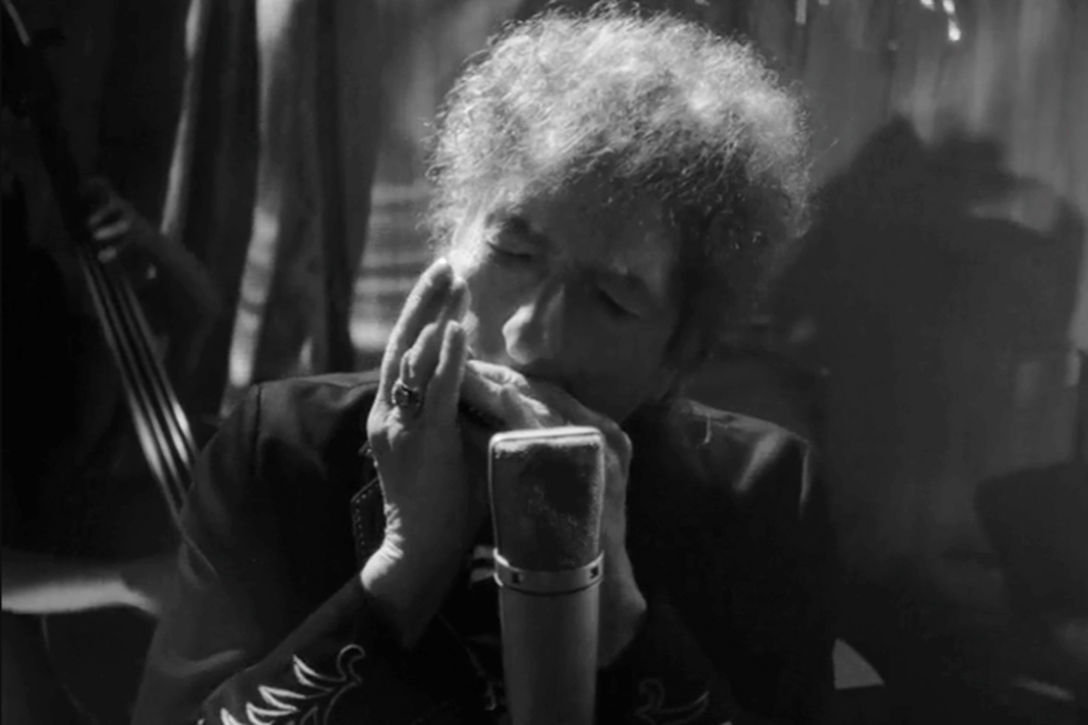 A black and white photo of Bob Dylan, with curly hair, playing a harmonica in front of a vintage-looking microphone. 