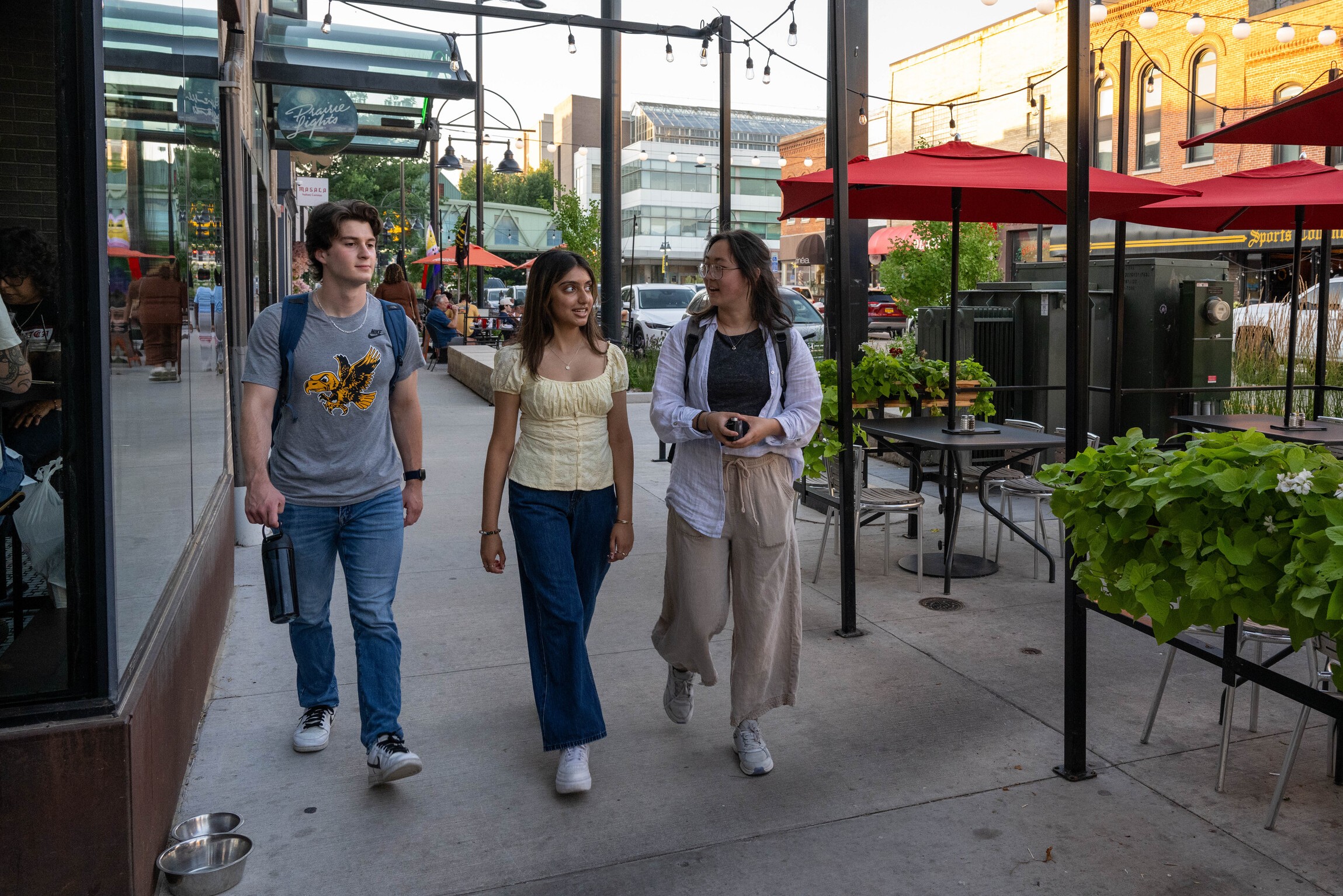 Three students walking cheerfully through a downtown Iowa City restaurant area, with outdoor seating to their left.