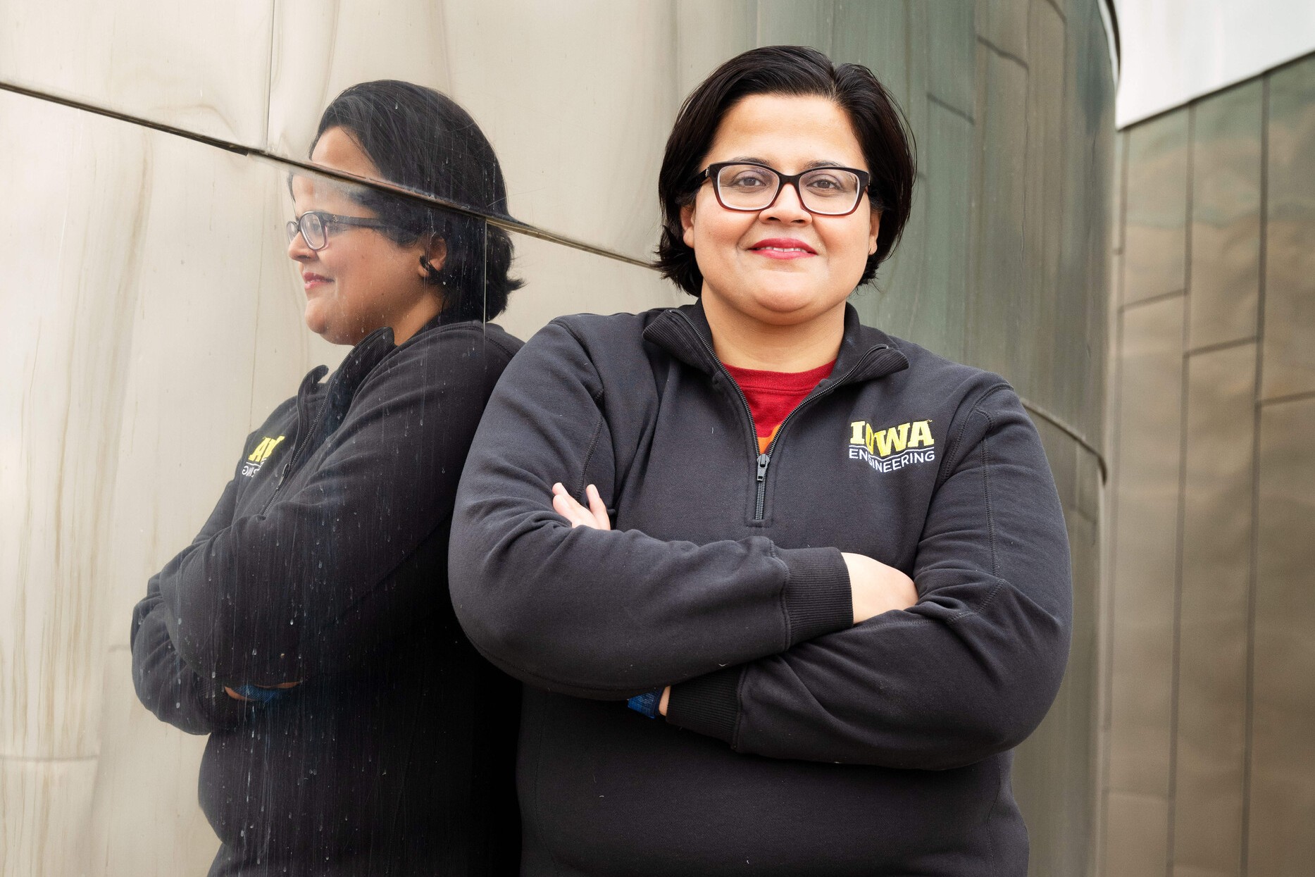 Fatima Toor, a woman with short black hair in a bob style, black glasses, a red shirt and a UI jacket. She's standing against a mirrored building and you can see her reflection behind her.