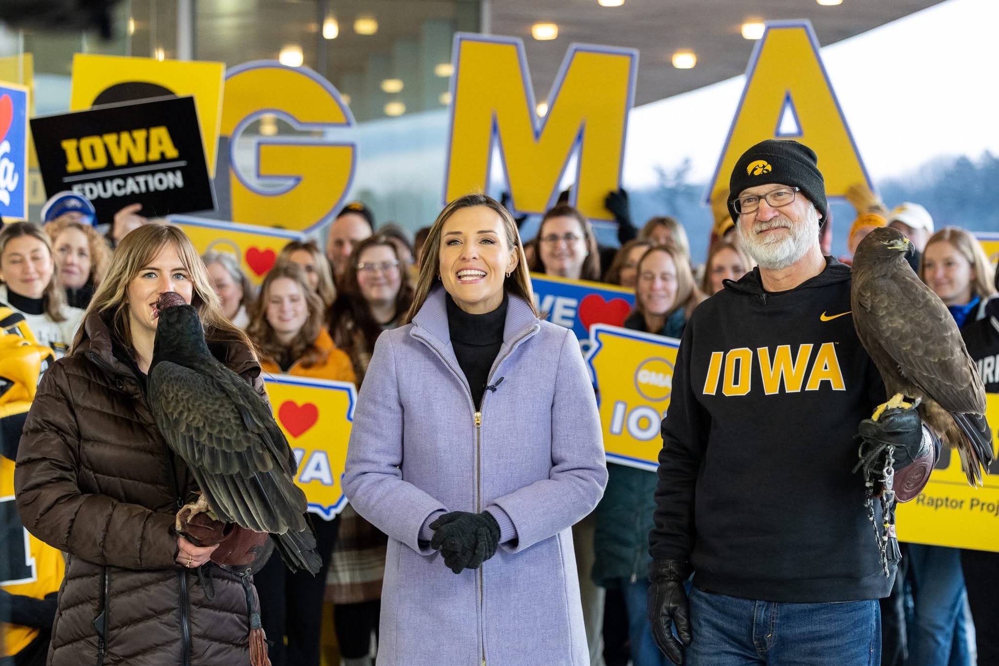 Staff from the Iowa Raptor Project holding hawks stand with Mary Bruce from ABC News. The crowd behind them are holding up a sign that reads GMA (for Good Morning America)