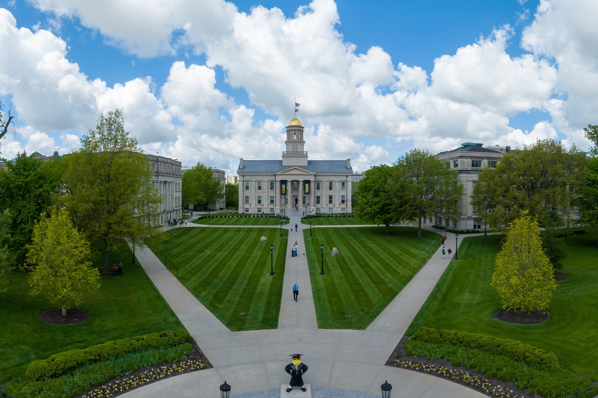 Paths cutting through the Pentacrest, leading to the Old Capitol building on a spring day. The sky is a vibrant blue with puffy, fair weather clouds.