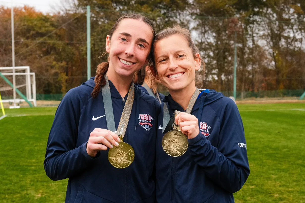 Paige Beaudry and soccer teammate standing on a soccer field holding Olympic gold medals 